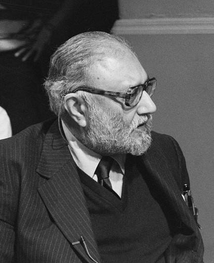 Photo en noir et blanc d’Abdus Salam portant un costume sombre à rayures, une chemise blanche et une cravate foncée. En arrière-plan, une autre personne est partiellement visible.