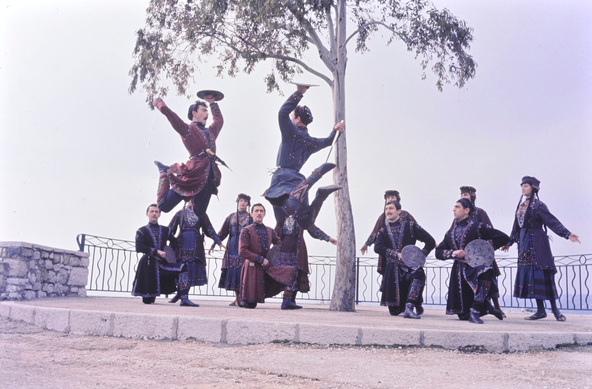 Paris, 1965. A group of twelve Georgian men and women in traditional dress. Four of the men are kneeling, and six of the women are standing behind them, all facing the spectacle in the center, where two men are engaging in a mock battle or sword dance. Both of them have leapt high into the air, with their feet pulled up behind their rears, for some 1.5 m clearance above the ground; they are facing each other, each with a short sword in the right hand and a buckler (small round shield) in the left, as if about to hit each other. One of the men is also shouting dramatically (the other’s face is not visible).