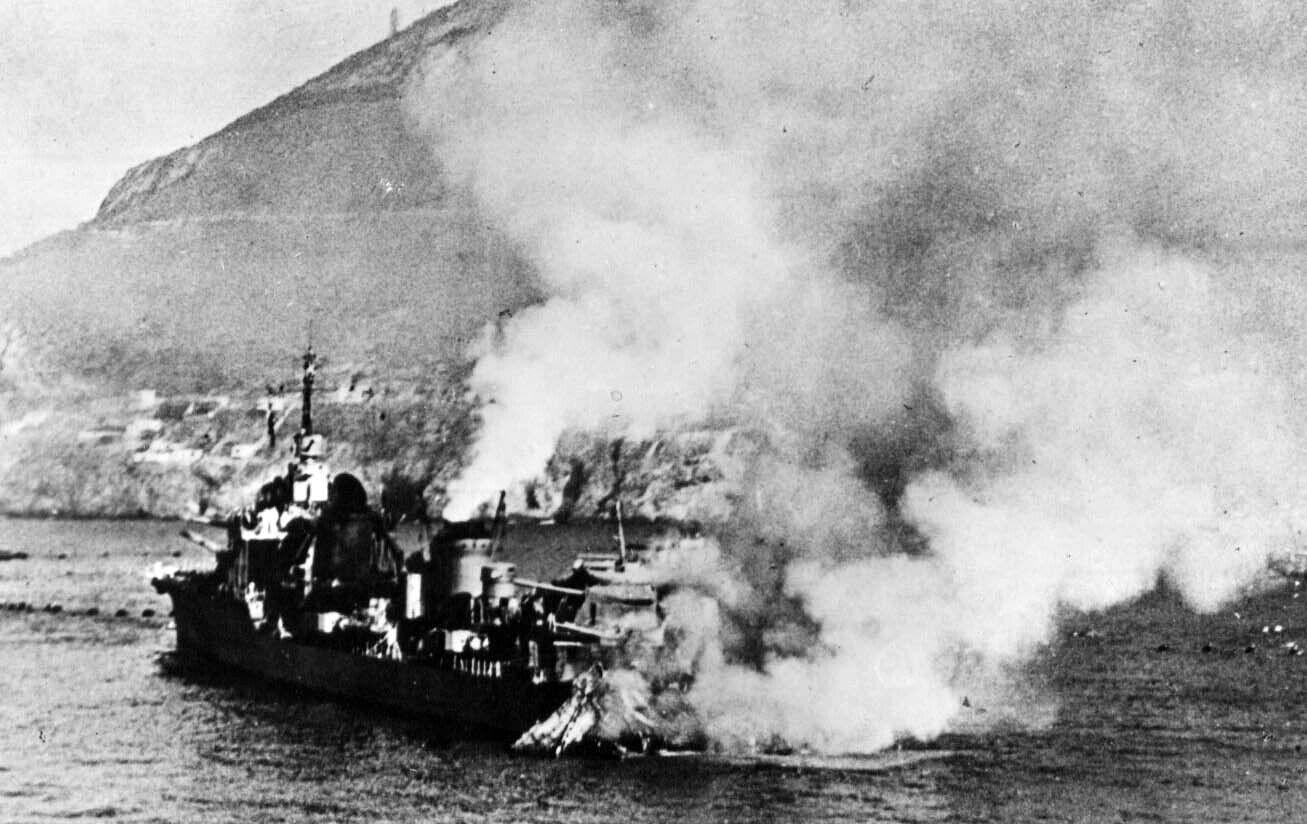 Une photographie granuleuse en noir et blanc datée de juillet 1940 du contre-torpilleur français Mogador en feu après avoir été touché par des obus de la Royal Navy lors de l'attaque de Mers el-Kébir. Le navire est visible en quart arrière gauche, naviguant vers la droite. Son arrière est en feu, avec d'épaisses colonnes de fumée noire et blanche s'élevant verticalement et se déplaçant vers la droite, obscurcissant une partie du navire. En arrière-plan, on distingue la colline escarpée et rocheuse dominant le port de Mers el-Kébir.