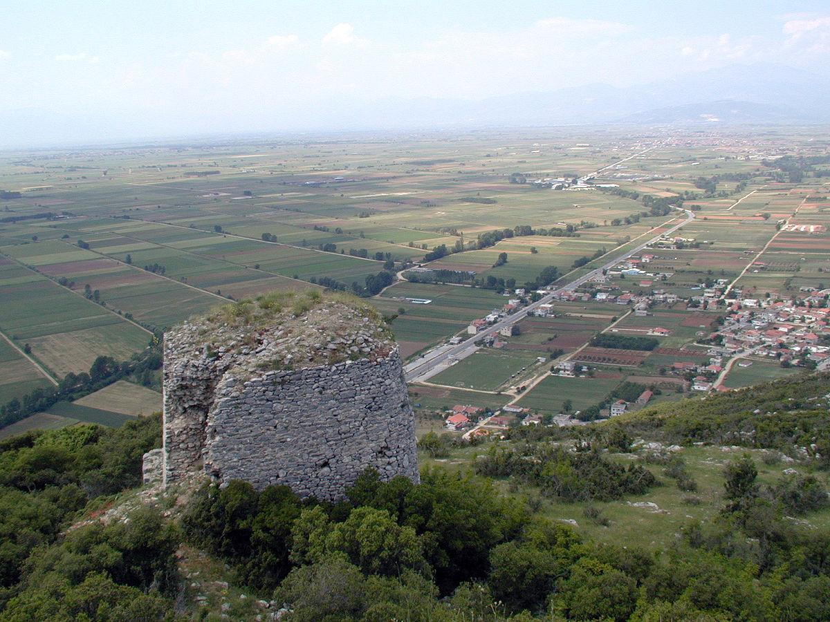 Une vue aérienne de la plaine de Philippes, dominée par une tour en ruine au premier plan, perchée sur une colline verdoyante. La plaine s'étend à l'horizon, parsemée de champs cultivés, de routes et de quelques habitations. Au loin, des montagnes se dessinent dans la brume. Cette vue est celle depuis l'acropole d'où Cassius observa la défaite de ses troupes lors de la première bataille de Philippes avant de se donner la mort.