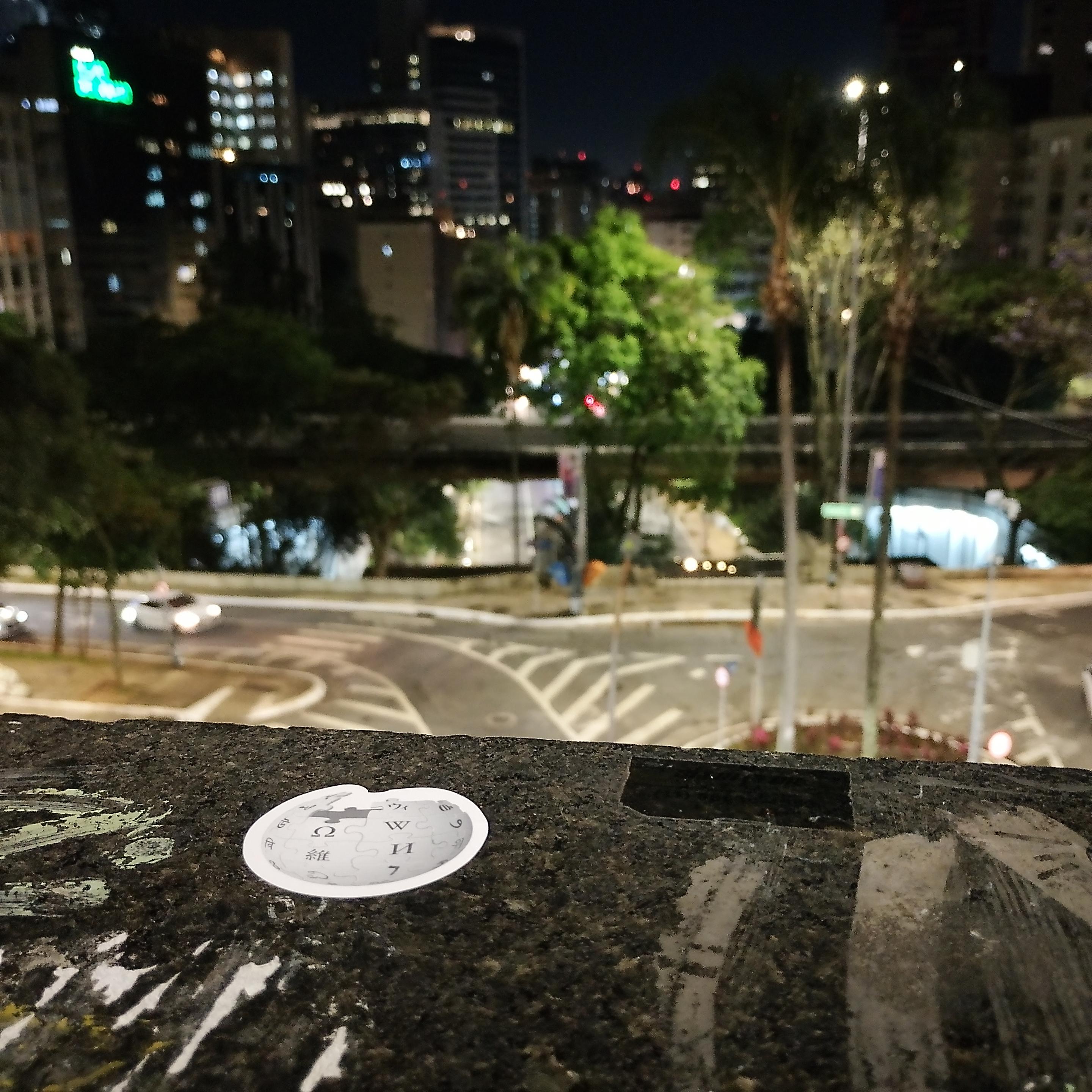 adesivo da wikipédia, em uma bancada, ao fundo São Paulo (atrás do MASP)