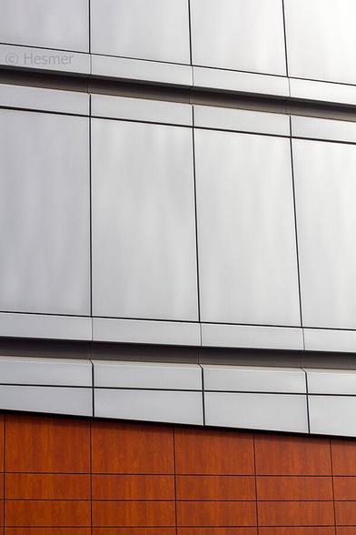 Die Fassade eines modernen Bürogebäudes. Unten rötliche Holzpanele, oben grössere Panele aus hellem Metall. Das Bild wirkt durch die unterschiedlichen Linien, die im Gitter verlaufen.