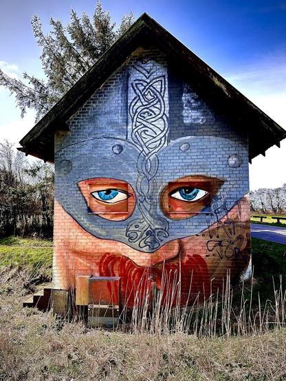 This image features a small building, possibly a utility or pump house, that has been creatively transformed with a mural to resemble a face. The artwork gives the illusion that the structure itself is a character, with eyes painted around two windows and intricate designs adorning the rest of the facade. The upper portion of the building is painted in dark tones with a grey pattern, reminiscent of Celtic or tribal designs, while the lower section simulates a beard in a red-orange hue. Additionally, there is graffiti on the left side of the building. The surrounding area includes trees, grass, and a clear sky above, contributing to the uniqueness of the scene.