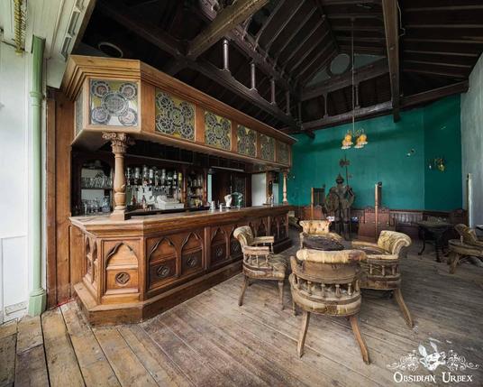 An abandoned Victorian bar with an ornate wooden counter, stained glass panels, and several worn chairs on a wooden floor. A knight armour stands near a teal wall.