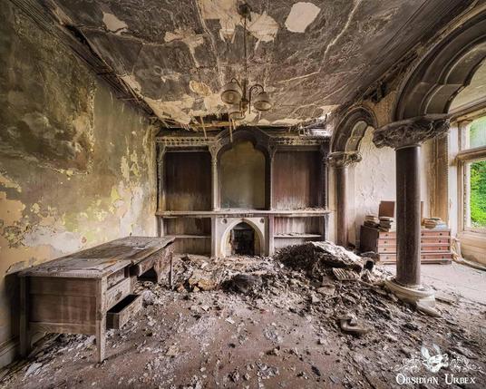 An ornate, fire-damaged room in a Victorian ruin, featuring peeling walls, a collapsed ceiling, and debris covering a desk and fireplace.