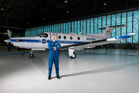 Pilot Jeremy Johnson stands in a large hangar building in front of a blue-and-white PC-12 propeller aircraft with his arms crossed. He is wearing a dark blue NASA flight suit and black boots. Visible on the side of the plane are NASA logos, text that says, “Glenn Research Center” and “N606A,” and a small American flag. Pilot Jeremy Johnson stands in a large hangar building in front of a blue-and-white PC-12 propeller aircraft with his arms crossed. He is wearing a dark blue NASA flight suit and black boots. Visible on the side of the plane are NASA logos, text that says, “Glenn Research Center” and “N606A,” and a small American flag.