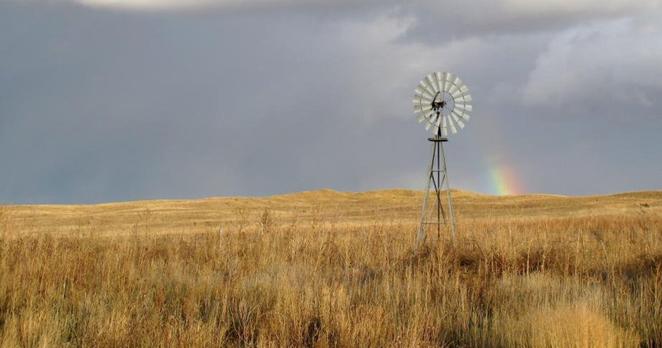 An old metal windmill stands in a field of brown grass. The sky is cloudy and there is a slight rainbow in it.