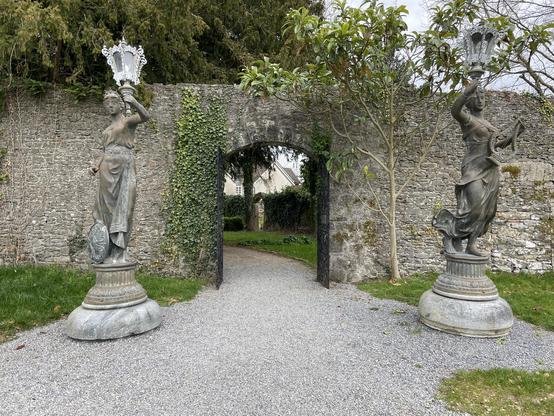 The opposite side of the stone wall, as described in the other photo. On this side there is less ivy, and both stone sculptures can be seen. A short tree is also growing on the right side of the opening.