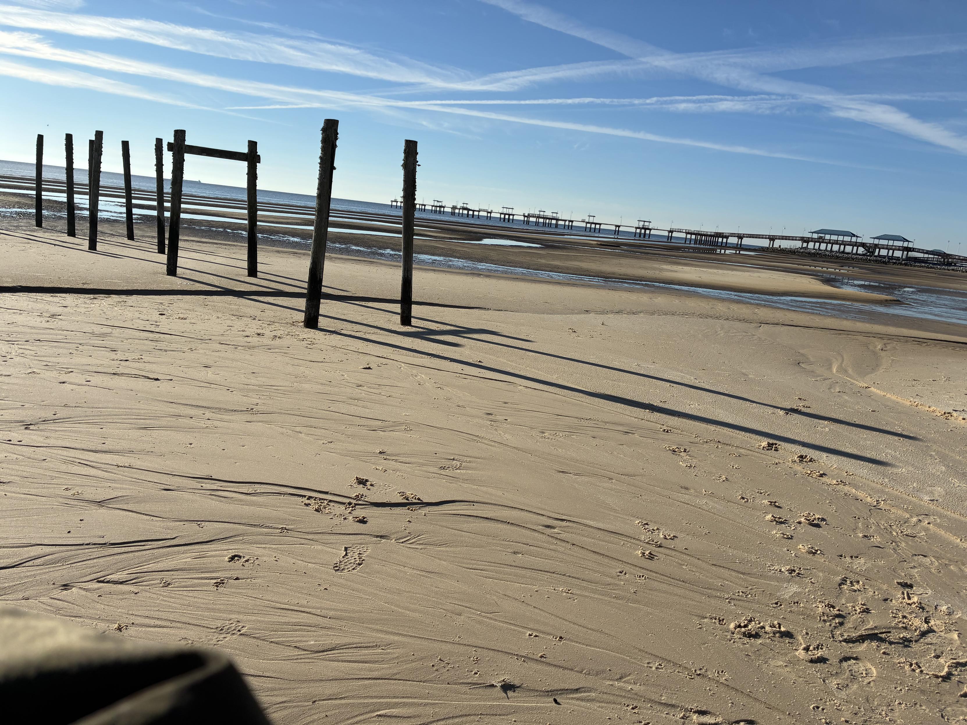 beach shot with water-sculpted sand in the foreground, abandoned pier remains casting shadows on the left, and clouds striating a blue sky
