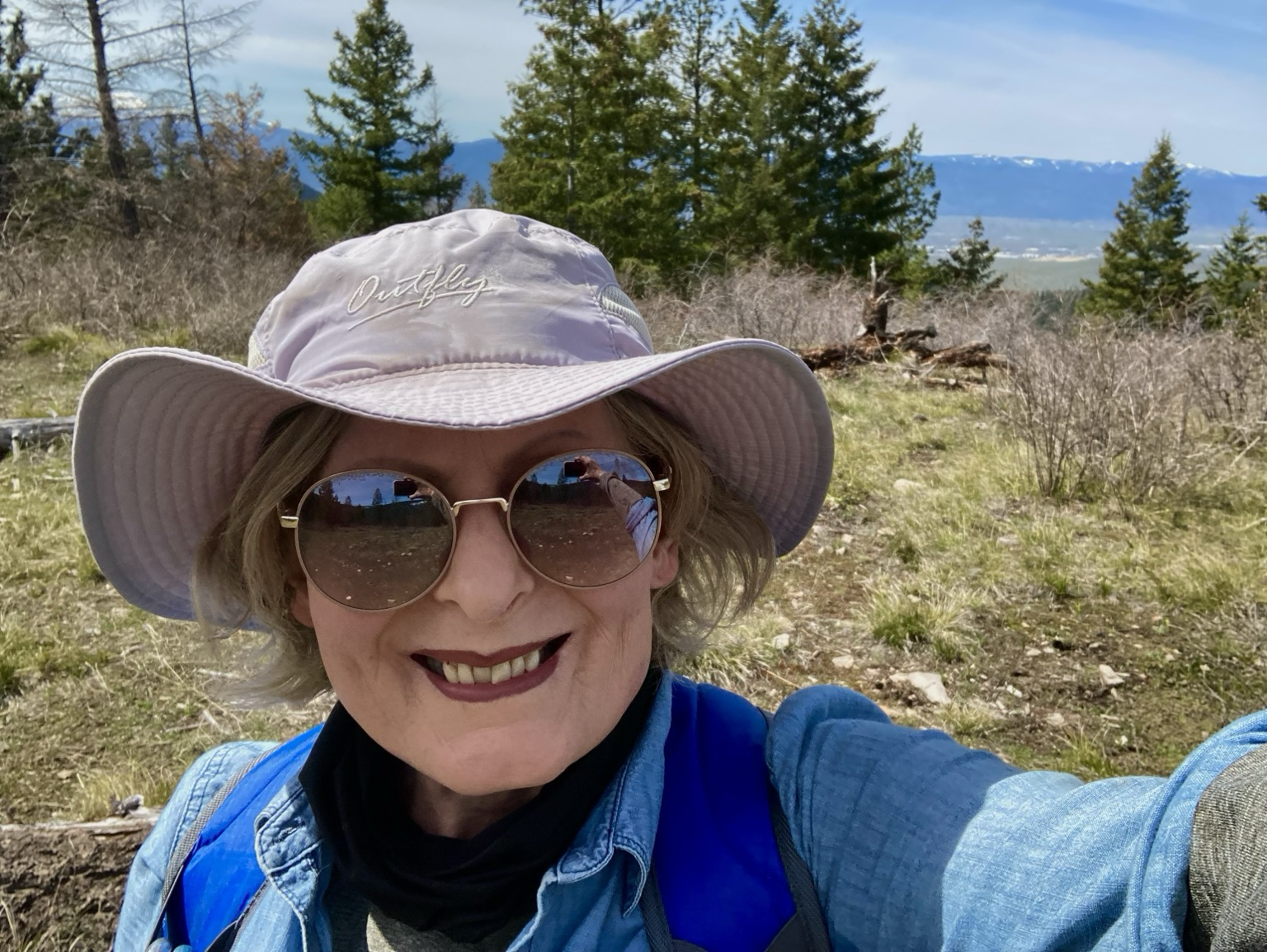An older blonde woman wearing sunglasses, a beige hat, and a blue shirt stands in a clearing in a forest, a valley and mountain range visible in the distance behind her. The sky is hazy.