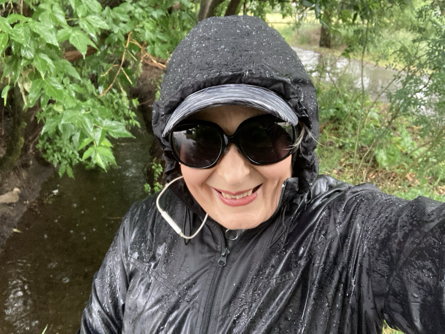 An older woman in a black rain jacket stands in the rain in front of trees, brush, and an irrigation ditch. She’s wearing sunglasses due to sensitive eyes.