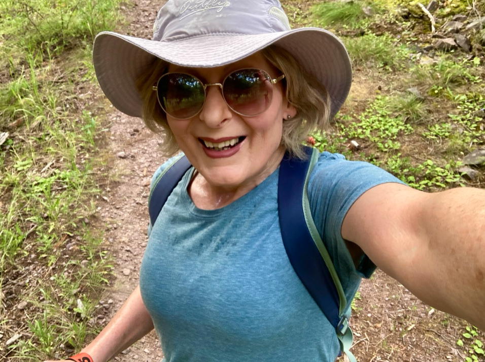 An older blonde woman stands on a forest trail surrounded by flora. She's smiling and wearing sunglasses, beige hat, and a blue top. The straps on her light pack are visible.