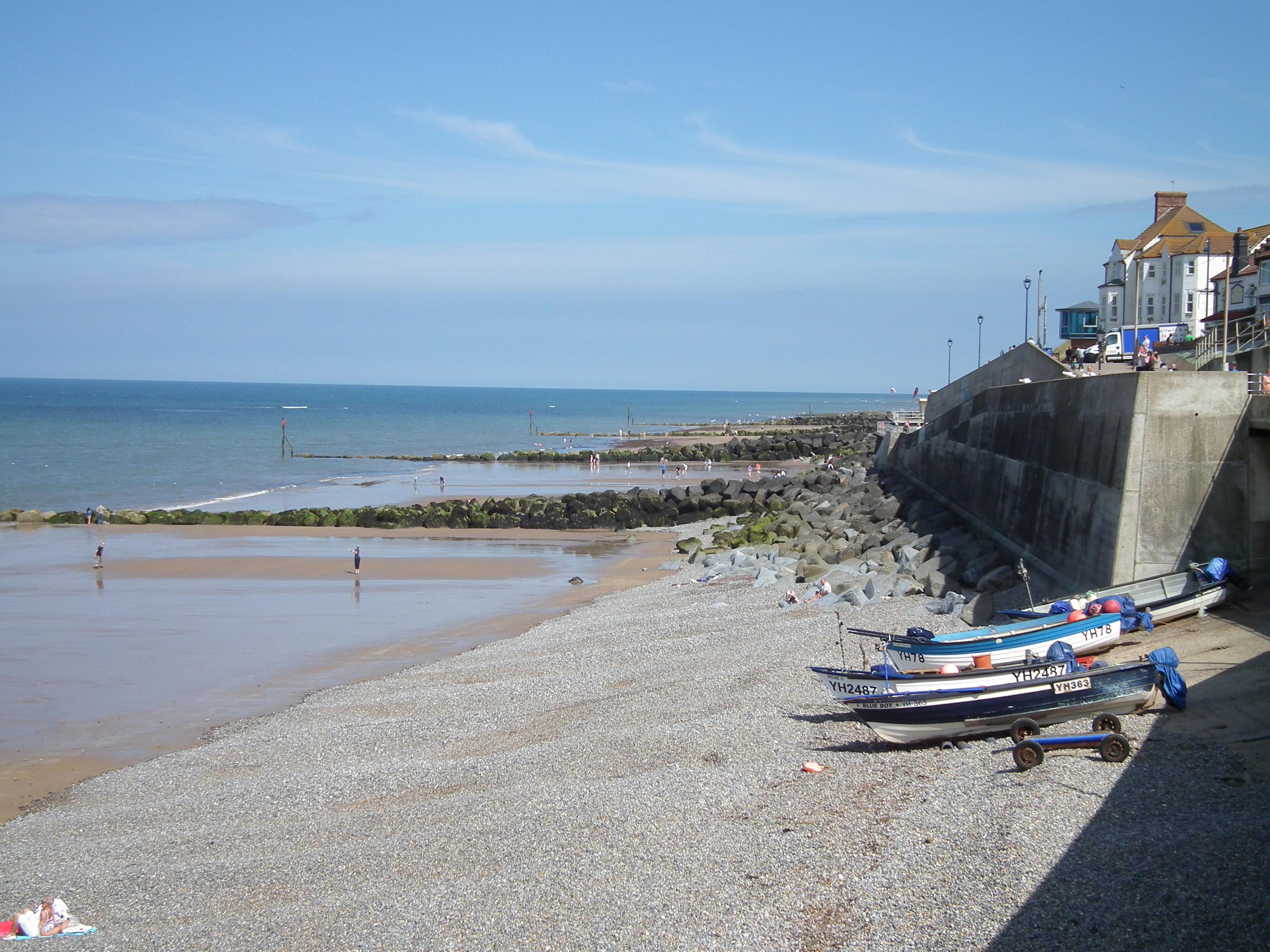 A view of the beach at Sheringham in Norfolk - the tide has retreated, exposing a wet sandy beach broken up by stone flood defences. A couple of traditional fishing boats are moored up at the top of the beach, next to a raised promenade with white coastal cottages on top.