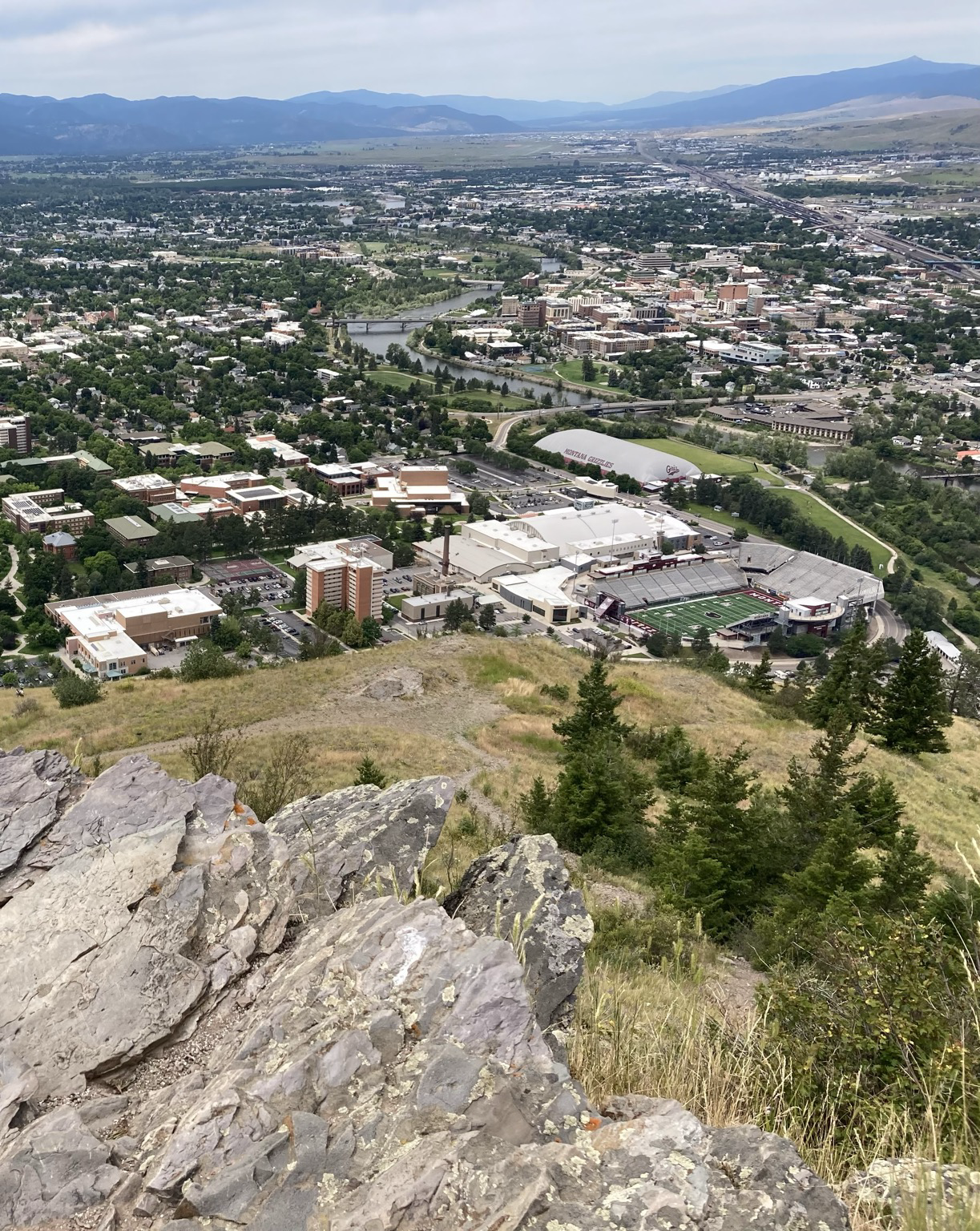 From a rocky perch on the side of a mountain, the view looks down a steep slope, a city spread out below. The view then extends across a broad valley to distant mountains.
