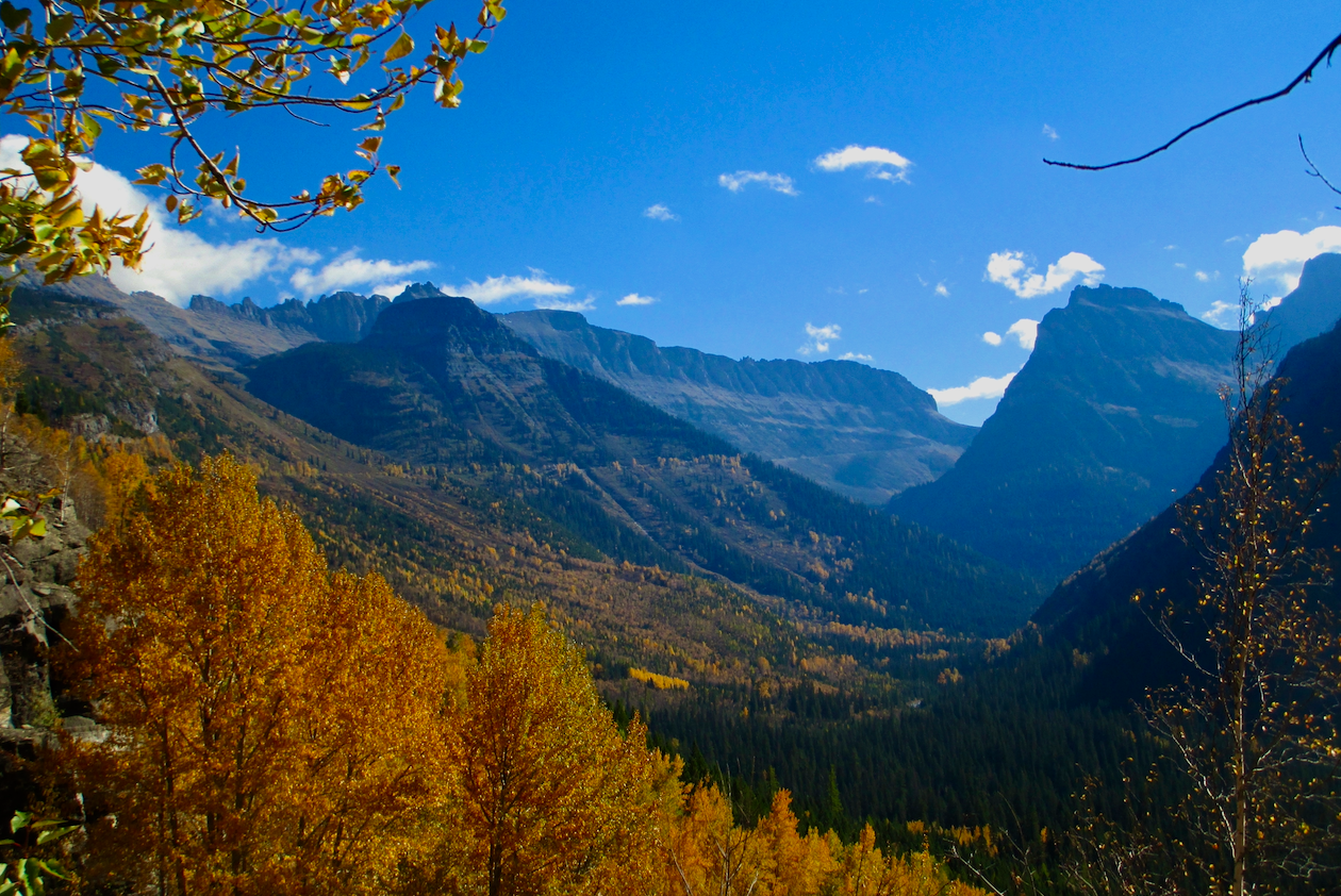 Taken in autumn, the view looks over trees adorned in red leaves, one with golden leaves lining the border on the left. Peaks rise from a forest of color in the background, the mountains silhouetted, the sky mostly clear.
