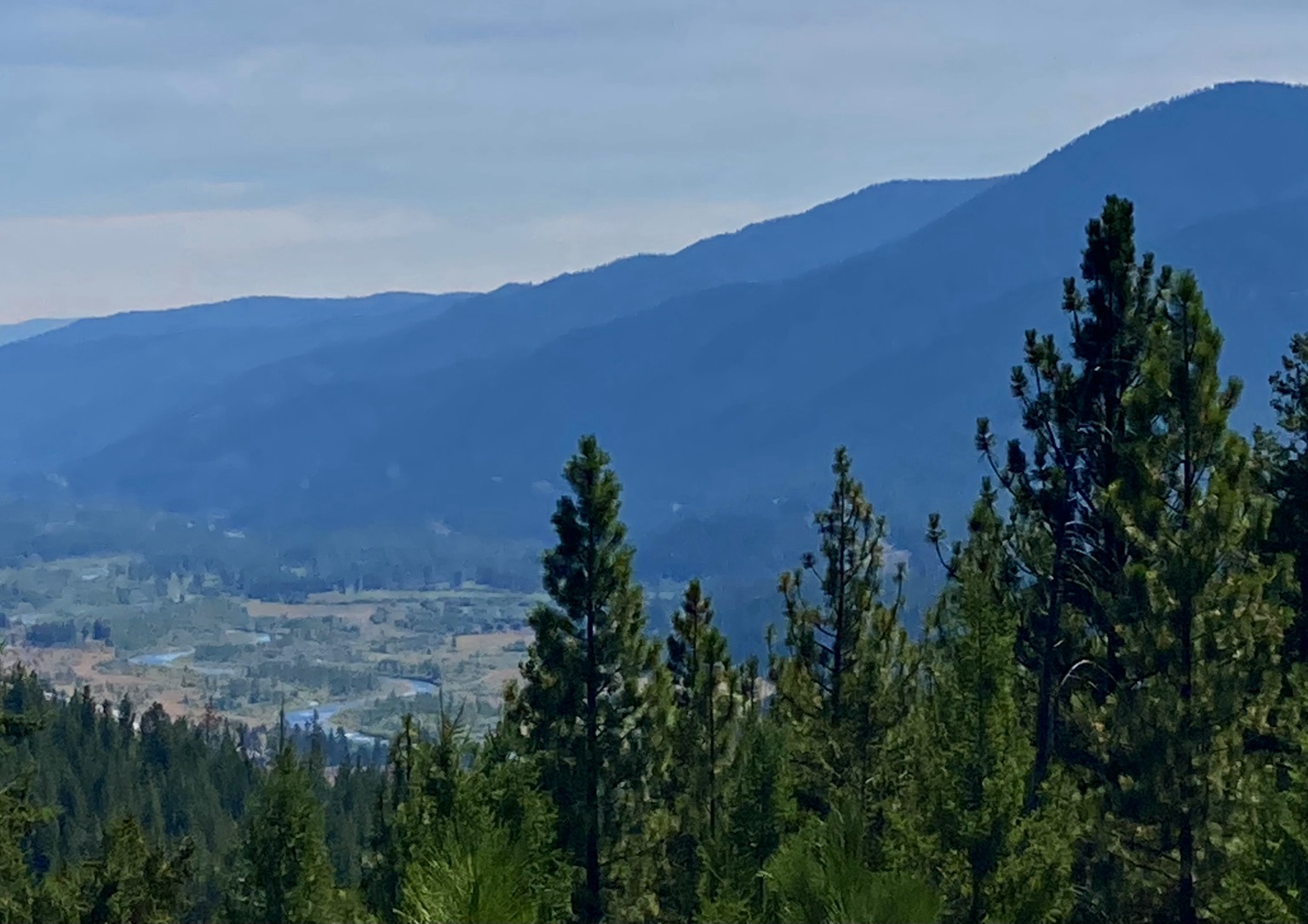 The few looks over pine forest and down a valley where a river flows, the mountains lining the valley appearing like layers. The sky is hazy.