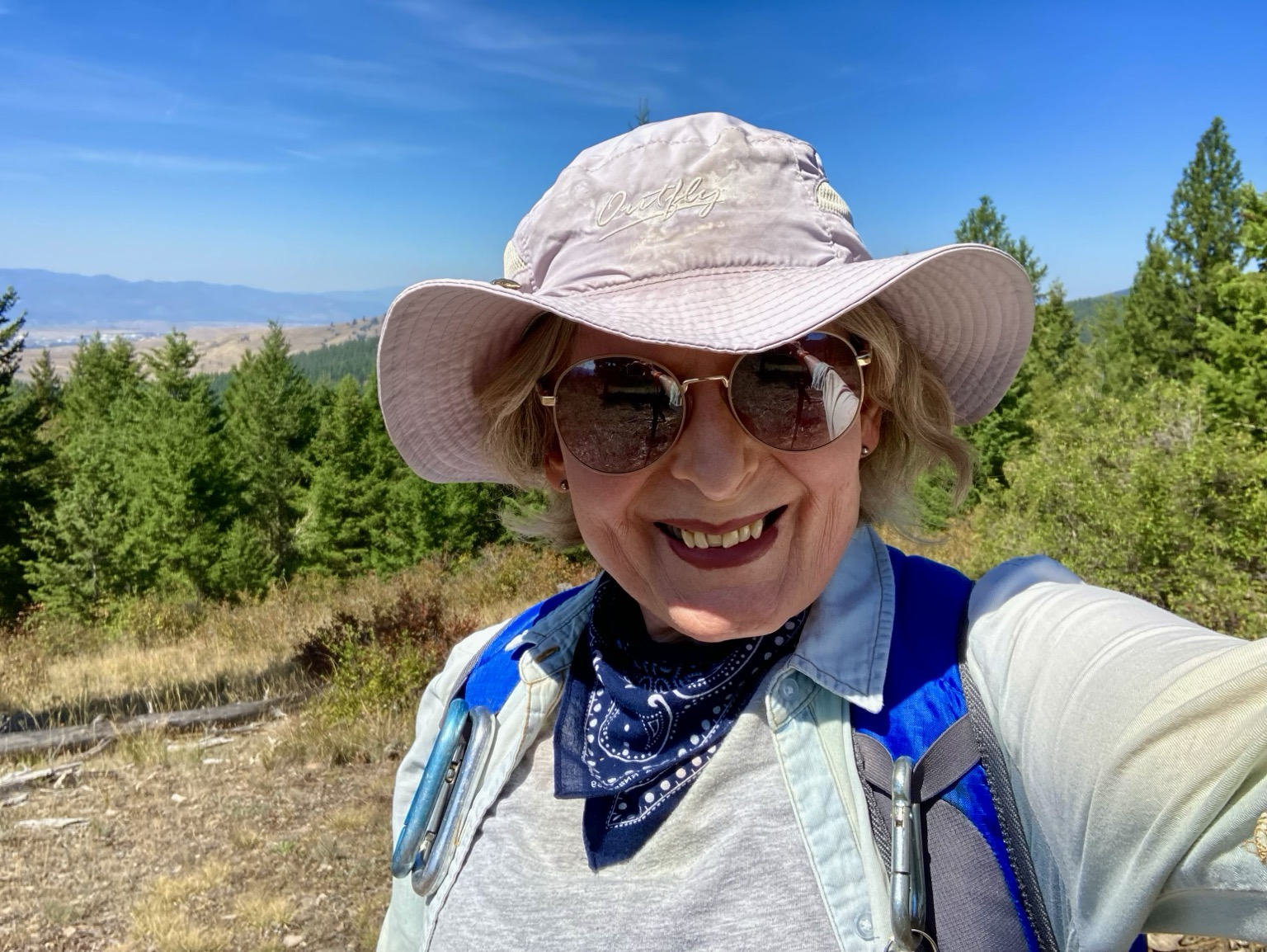 An older blonde woman wearing a light hat, sunglasses, blue bandanna, and light blue shirt stands atop Milltower Ridge.