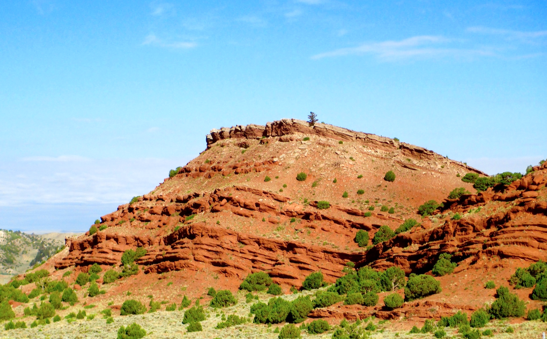 A red bluff dotted with green trees rises in central Wyoming, its layers clearly visible. The sky is clear, but hazy.