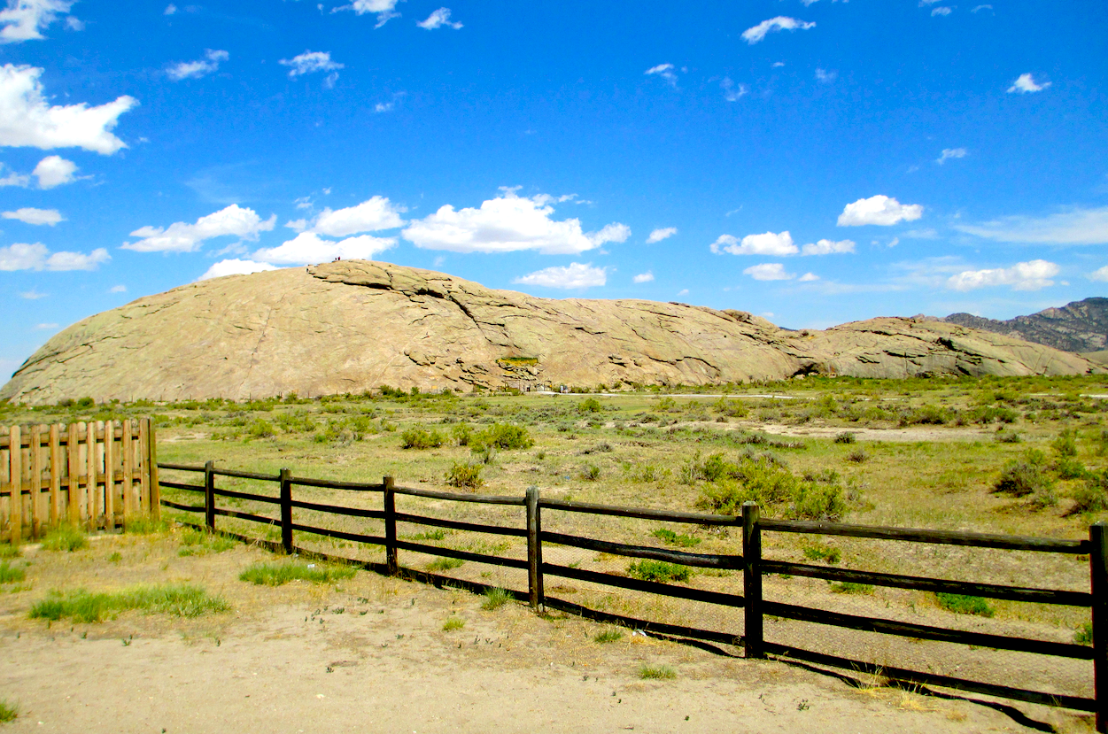 Beyond a wooden fence on the prairie, and beneath a partly cloudy sky, rises Independence Rock in Wyoming, a mound of solid rock that stands alone.