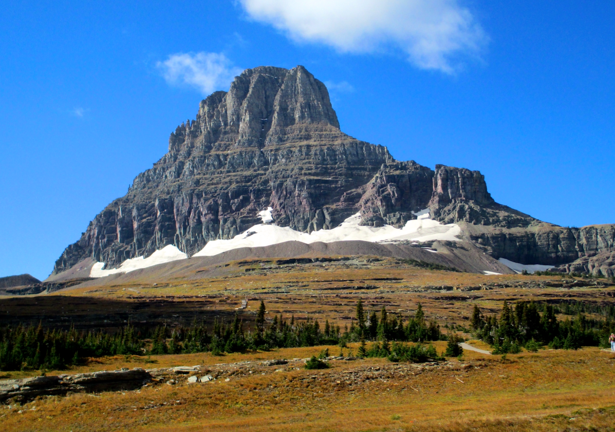 A rocky promontory in Glacier National Park in Montana rises beneath a mostly clear sky, the grass surrounding it a deep gold. The layers of sedimentary rock are easily visible, and there's snow around its base.