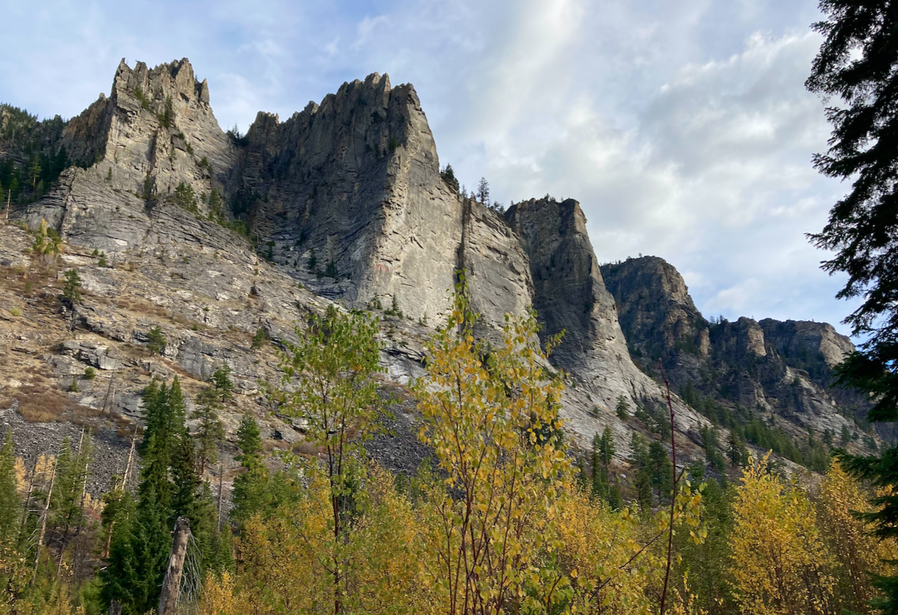 High above trees with golden leaves mixed with pines rises a wall of rock that becomes separate prominences beneath a cloudy sky.