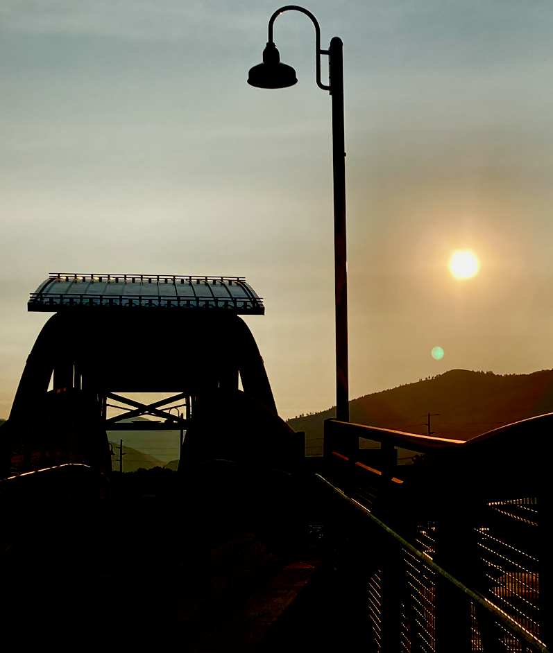 A bridge structure, light pole, and distant mountain are all silhouetted in the morning sun that is hidden behind a smoky sky.
