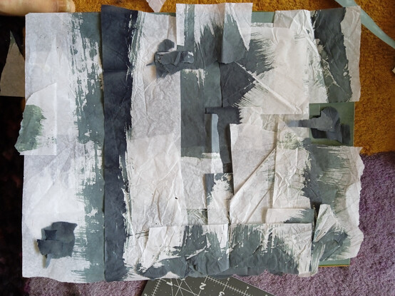 Close up of overlaid strips of white tissue paper with brush marks in grey-greenish blue, laid in an abstract pattern on a rectangular base.