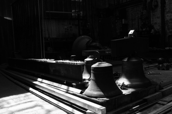 Black and white image of a row of bells stored on the workshop floor of the bellfoundry.  Play with diagonal lines and shadows.