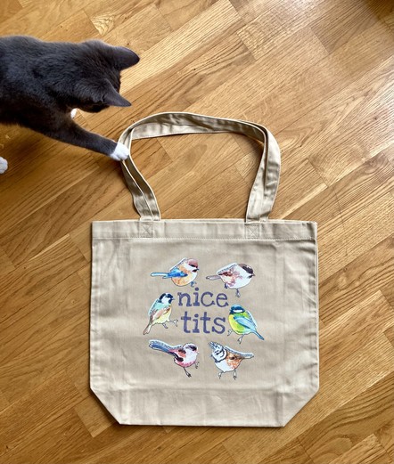 Ochre coloured cotton tote bag laid out flat on a light wood floor. The tote bag side facing the camera has six illustrated song birds, known as tits, on it along with the words "nice tits". A small grey kitten is coming in from the upper left corner of the photograph to paw at the tote bag handles. 
