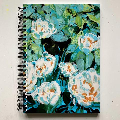 An overhead view of a spiral notebook on an off-white table top. The metal spiral is black and on the left. The cover has printed on it a section of an abstract painting of white tulips on a teal and green background of leaves. The tulip petals have shades of teal blue in them, and orange stamens.