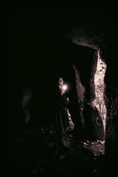 Monochrome photo of a person inside a cave holding their phone up shining the torch in the direction of the camera; the opening of the cave is visible on the right.