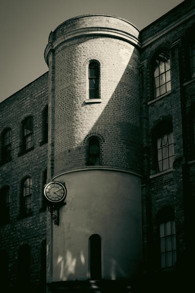 An old brick building with a round tower on which is a clock; strong shadows.
