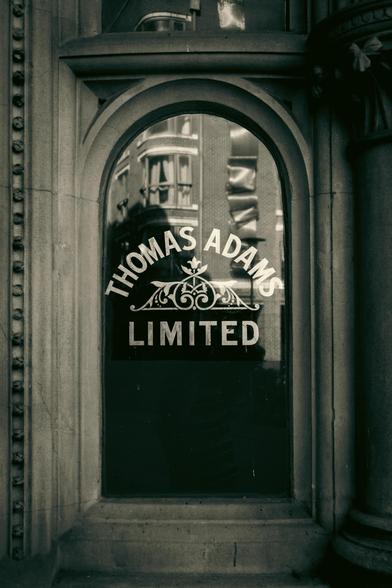 Window with the text: "Thomas Adams Limited" and an ornamental device.  The buildings opposite are reflect in the window.