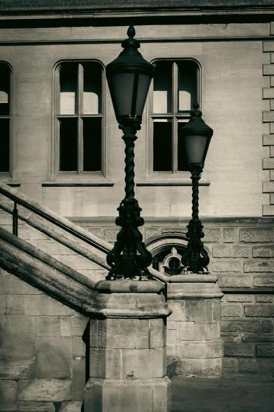 Two lampposts on top of stone pillars at the bottom of the stairs on the exterior of a building.