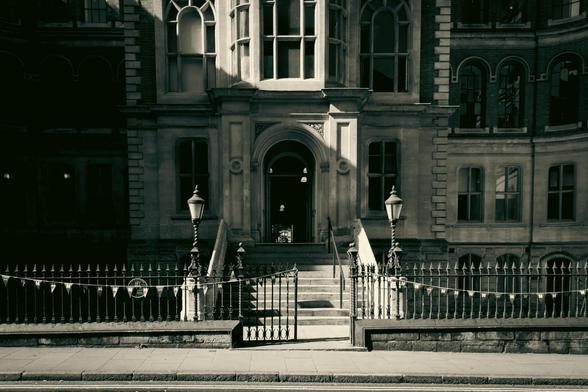 View from across the street at the front of a building; visible are iron railings and two lampposts flanking the stairs leading to the main entrance; heavy shadows.