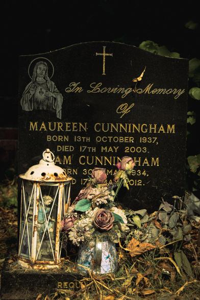 A rusty white lantern with a figuring inside and some withered flowers in a can in front of Maureen Cunningham's gravestone. She was born 13 October 1937 and died 17 May 2003.