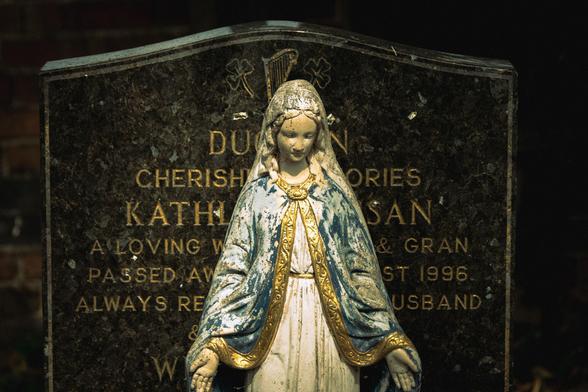 Statue of Virgin Mary in front of a black marble gravestone; the statue obscures the text.