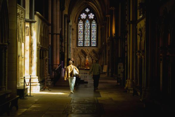 Interior of a goth cathedral; a patch of sunlight; two people walking towards a stained-glass window.
