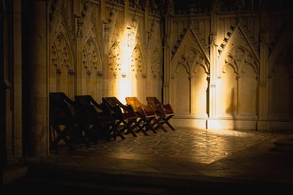 Inside a gothic cathedral: a row of wooden chairs against blind, arched windows, with patches of sunlight