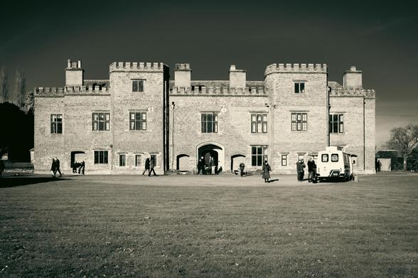 View across the lawn of a stately manor in the sunlight; people walking past; an ice-cream van is parked on the right.