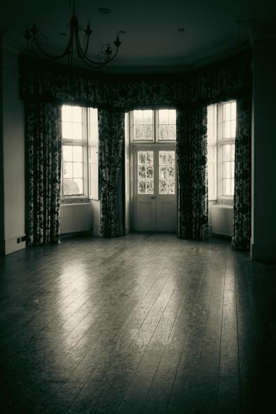 Inside a large room. A wooden floor, reflecting the light, looking towards a large bay window and door.