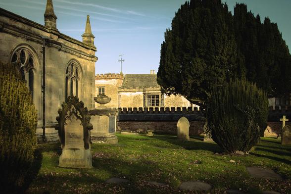 Side of an old church, gravestones and some trees; the manor house visible in the back.