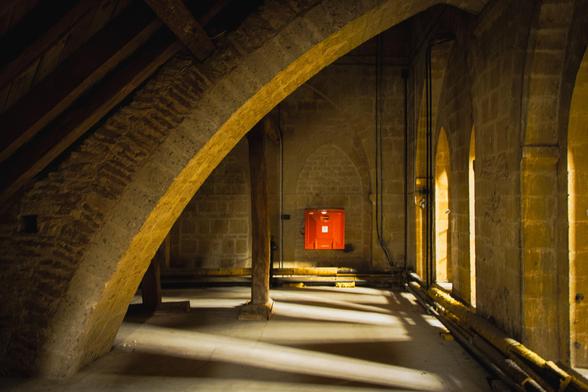 Upper gallery underneath the Cathedral's interior arches; on the right arched windows from where light floods in; on the back wall, red fire safety equipment. 