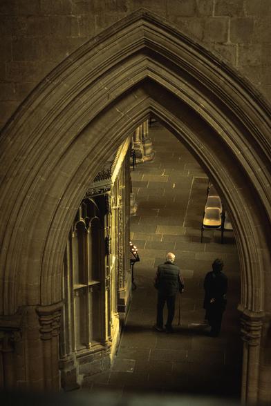 Cathedral interior: looking from above through an arched passageway; below are two people walking away.