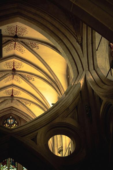 A section of the decorated, vaulted ceiling of the cathedral, bathing in light, seen through the openings of the Wells's famous scissor arches.