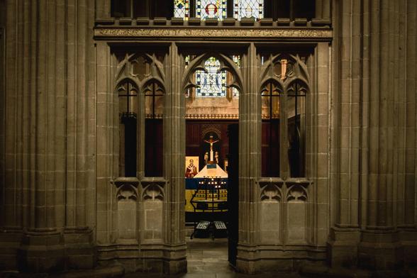 Interior of Wells Cathedrals: two stone pillars and a stone screen with arched windows and a opening that leads to the chapel behind in which we see an altar and a stained-glass window.