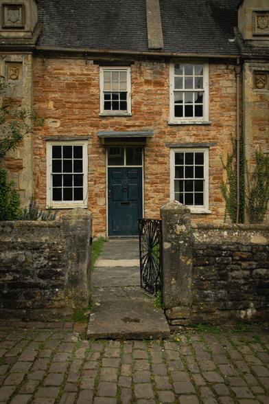 Stone wall with gate leading to the blue front door of an old house with four windows.