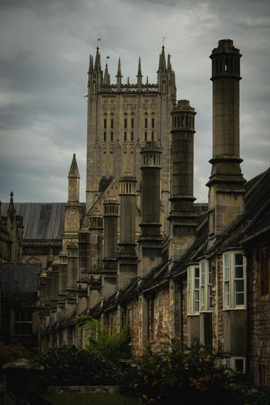 A row of old stone houses with high chimney stacks; ion the background the main bell tower of Wells Cathedral.