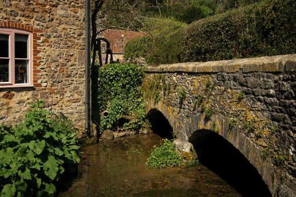 A stone bridge across the road next to an old cottage.  The bridge has two arches and the stones are covered in lichen.