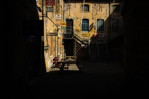 Courtyard of an old industrial building in the sunlight with deep shadows.  Vintage branded signs decorate the walls. A staircase leads to a door on the first floor.